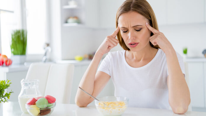 Photo of pretty housewife holding temples suffering terrible pain can't eat milk breakfast cornflakes have hangover after night party sitting table white light kitchen indoors